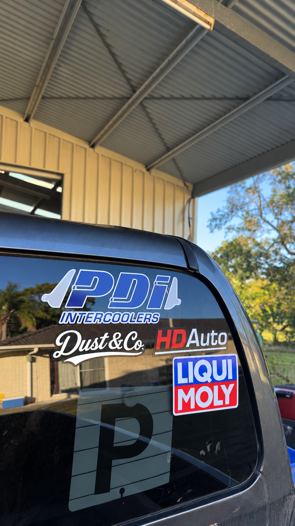A close-up of the rear window of a dark vehicle under a metal carport, featuring several automotive decals including PDI Intercoolers, Dust & Co. HD Auto, and Liqui Moly.