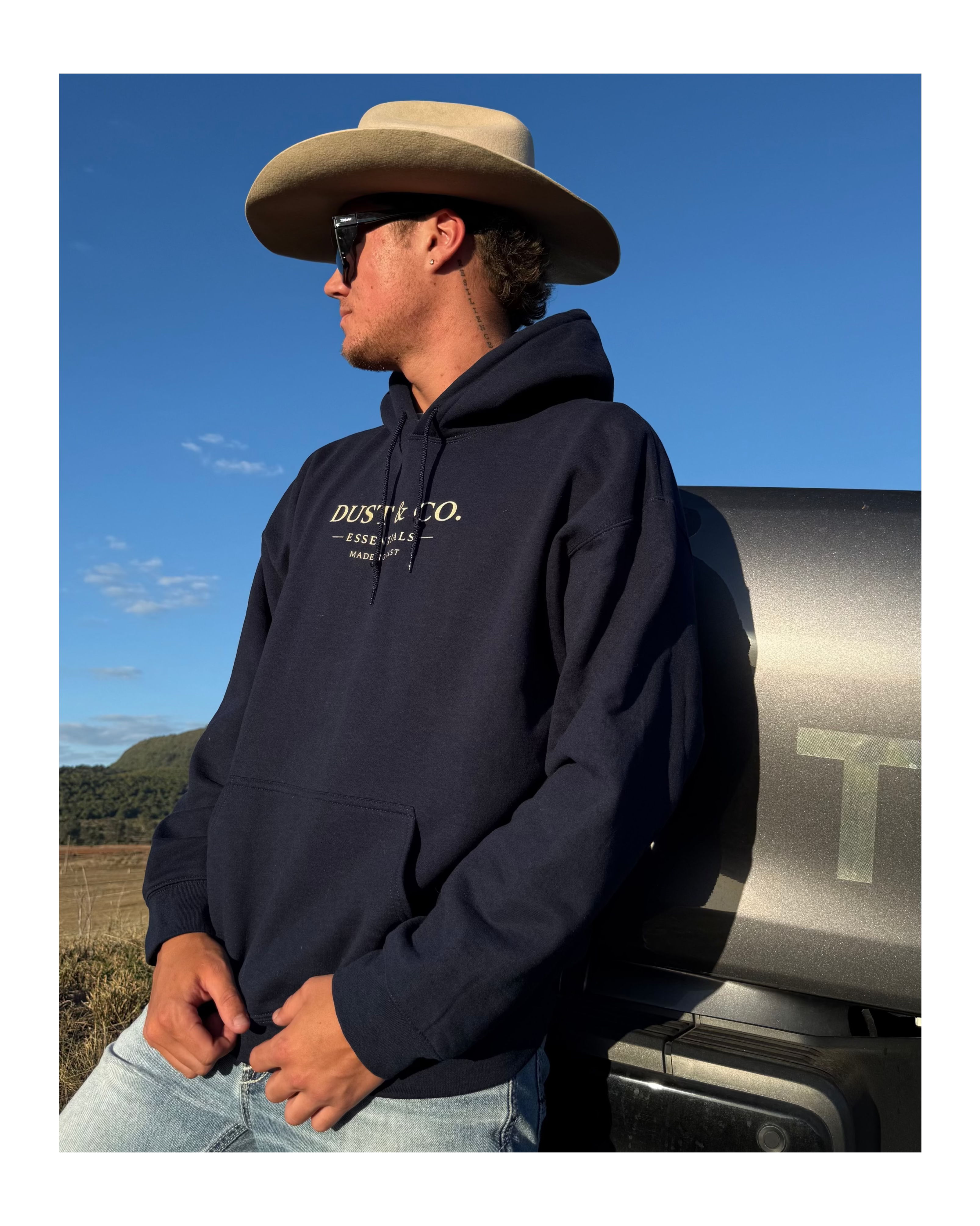 Man wearing a navy hoodie with 'Dust Co.' logo, cowboy hat, and sunglasses, standing next to a truck.