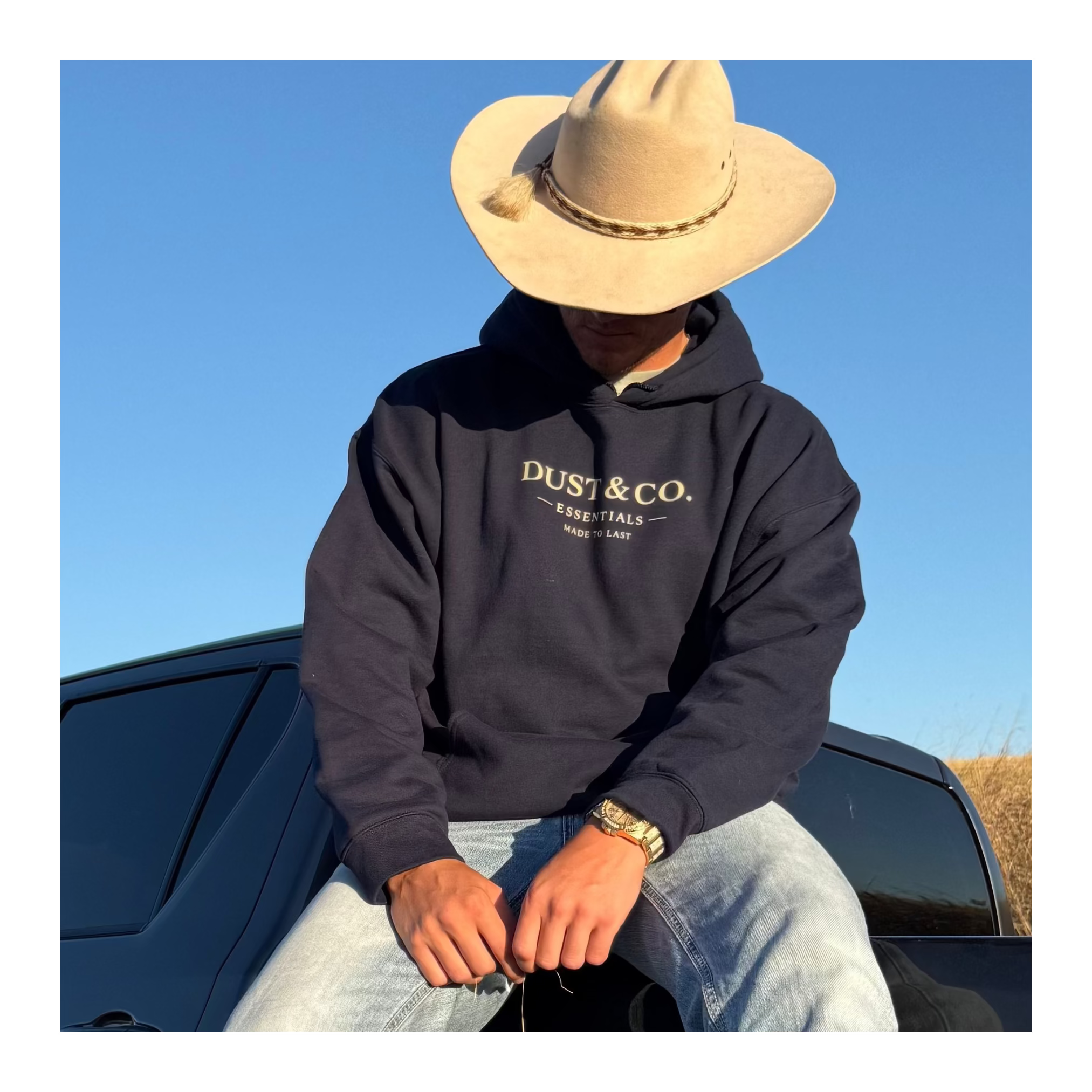 Person wearing a cowboy hat sitting on a black truck in an open field with clear blue sky.